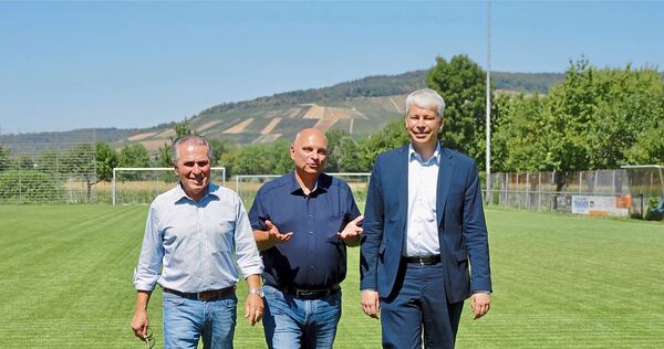 Bundesverkehrsminister Steffen Bilger (von rechts), Bürgermeister Jürgen Scholz und Umweltexperte Claus-Peter Hutter spazieren ü Bundesverkehrsminister Steffen Bilger (von rechts), Bürgermeister Jürgen Scholz und Umweltexperte Claus-Peter Hutter spazieren über den grünen, mit Mineralwasser gegossenen Sportplatz. Im Hintergrund sind vertrocknete und brachliegende Weinbergfläche