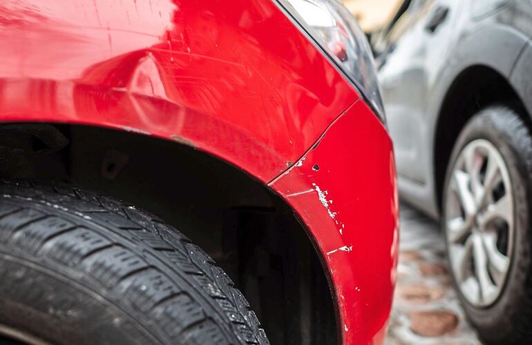 Das rote Auto steht mit einem Blechschaden in einer Parklücke, während ein graues Auto vorbeifährt. Die Regionalklasse bildet di Das rote Auto steht mit einem Blechschaden in einer Parklücke, während ein graues Auto vorbeifährt. Die Regionalklasse bildet die Schadensbilanz eines Zulassungsbezirks ab.